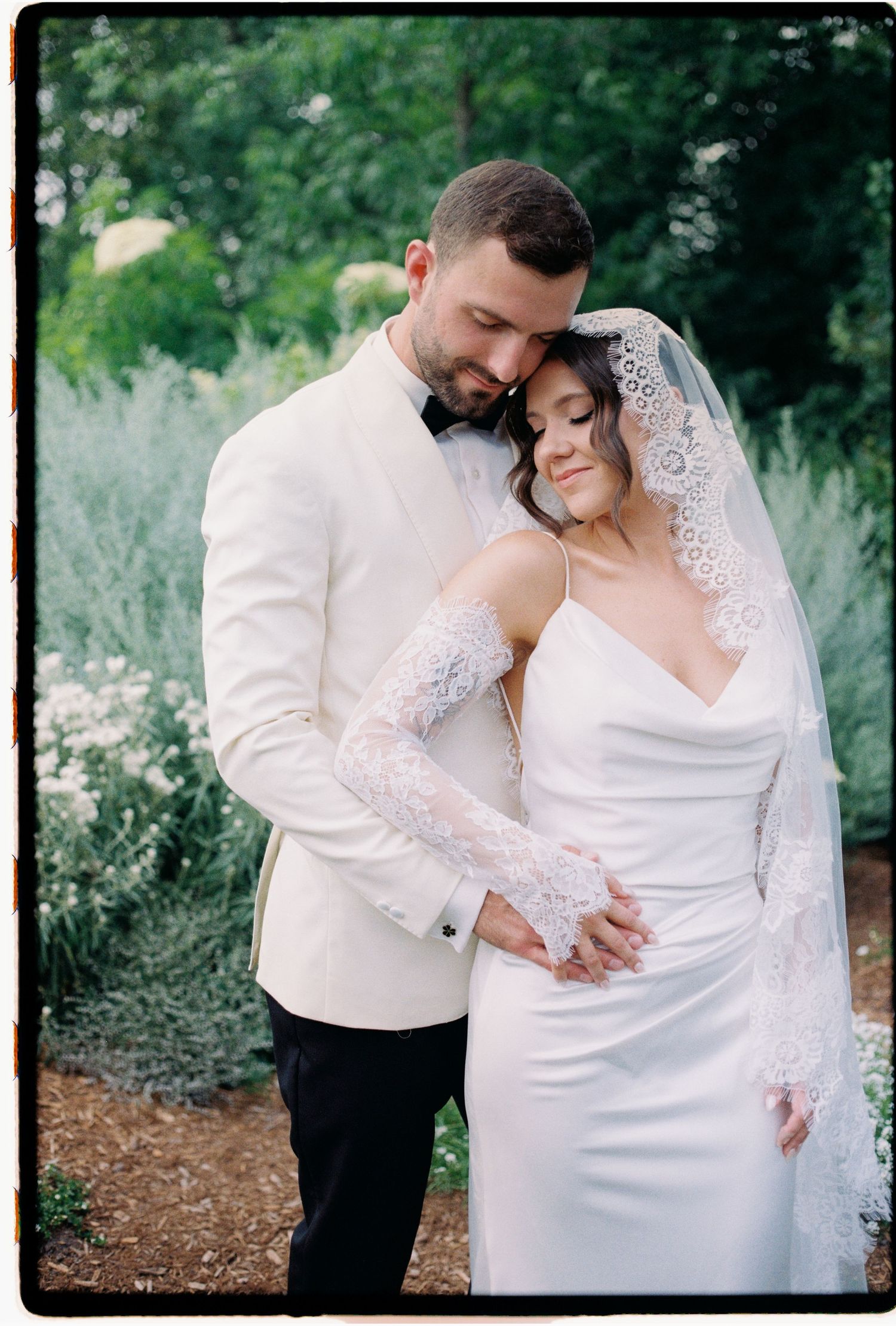 A couple shares an intimate moment in their wedding attire against a natural outdoor backdrop with greenery.