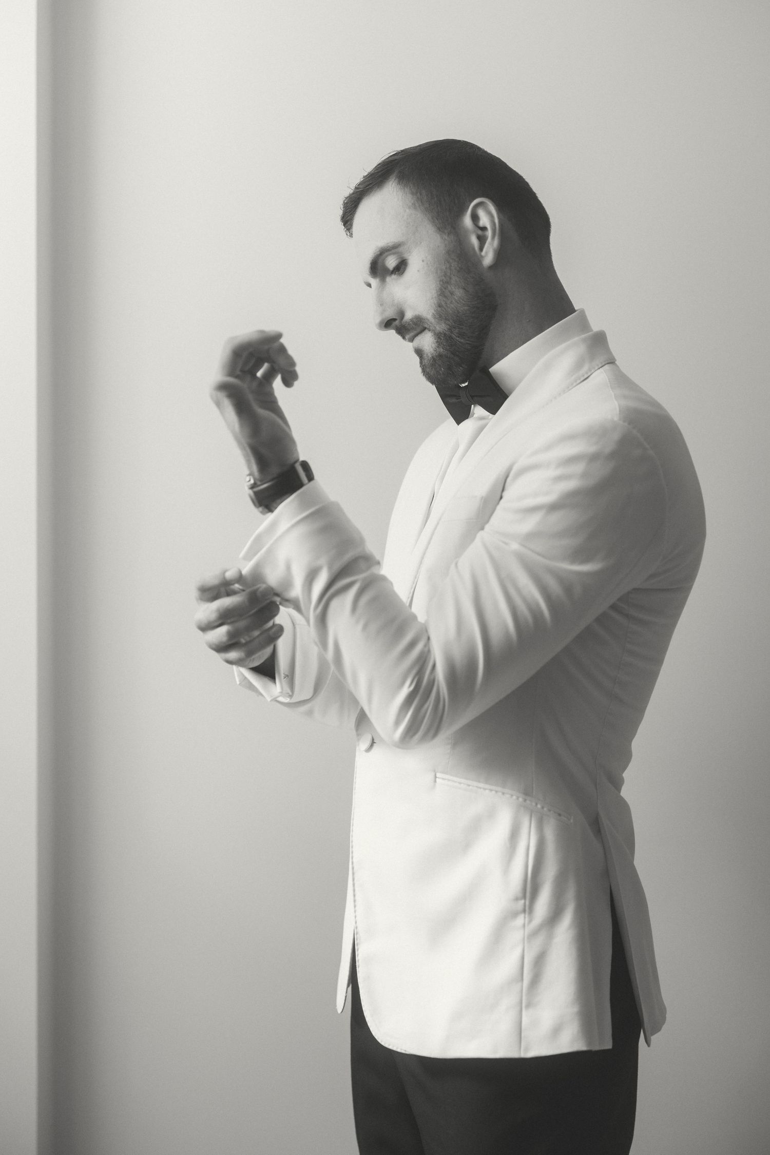 Side profile of a person in a white blazer adjusting their cuff against a light gray background.