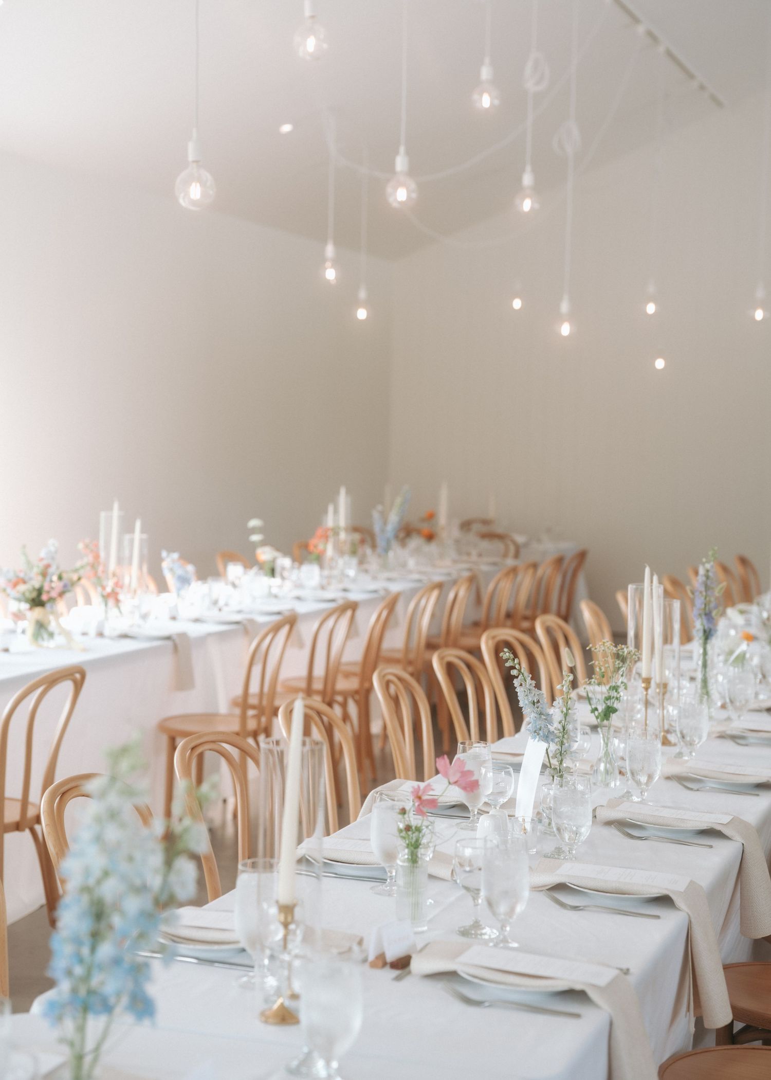 Elegant wedding reception table setup with white linens, bentwood chairs and hanging string lights in a bright modern space.