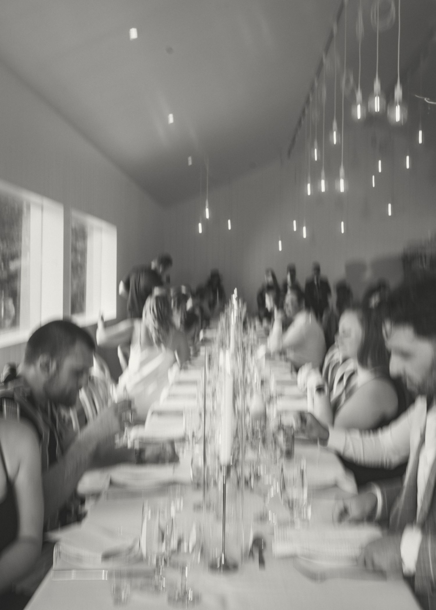 A black and white photo of people dining at a long table under modern pendant lights in a minimalist space.