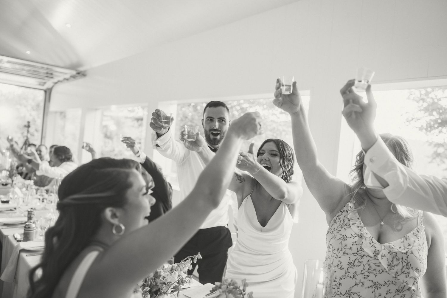 A joyful celebration scene at a wedding reception with people dancing and raising their hands under a white tent.