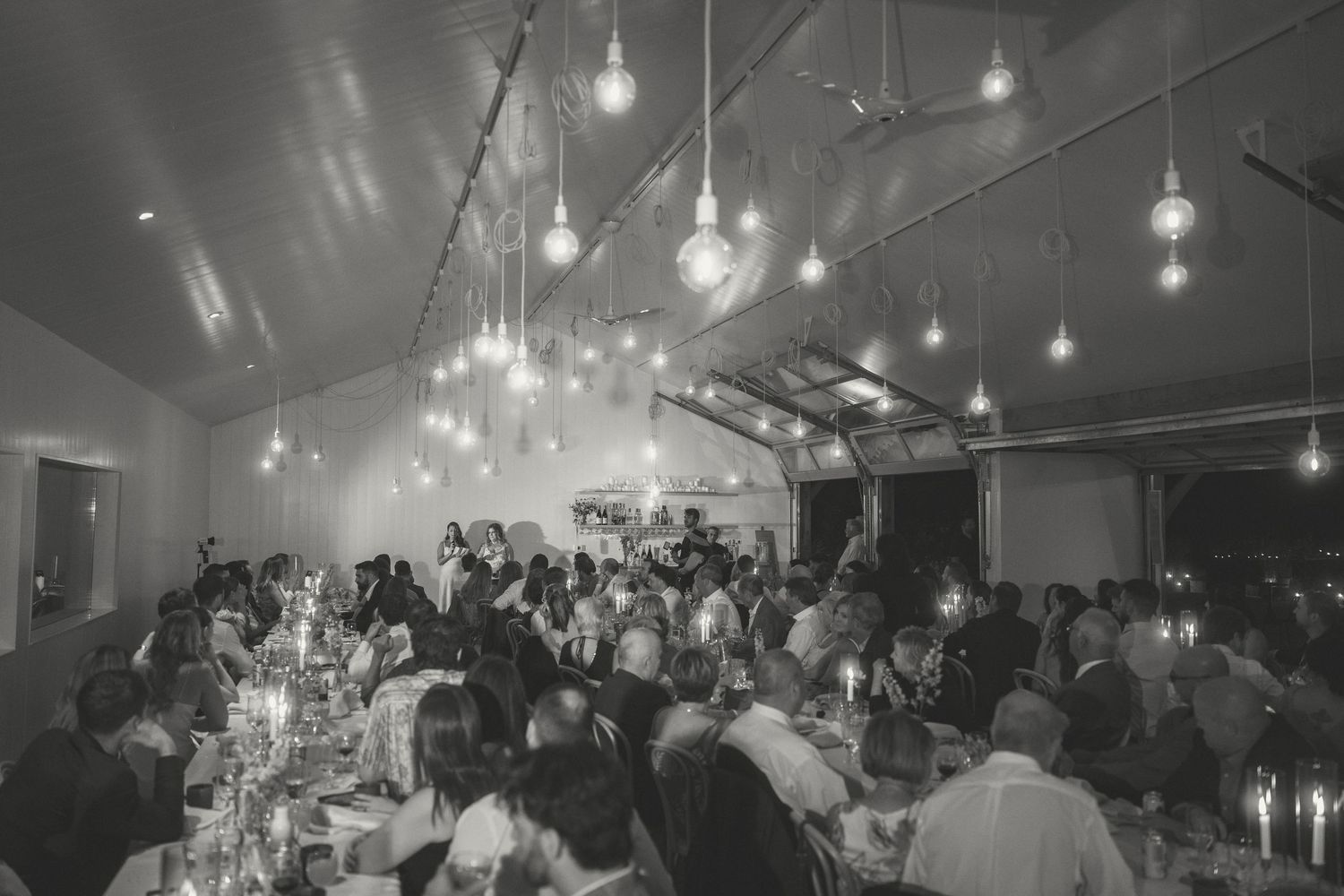 A crowded indoor event space with string lights hanging from the ceiling illuminating groups of people at tables.