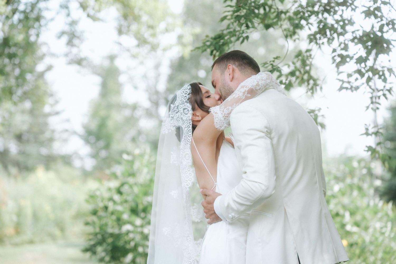 A romantic wedding couple shares tender kisses in a sunlit garden setting with soft greenery in the background.
