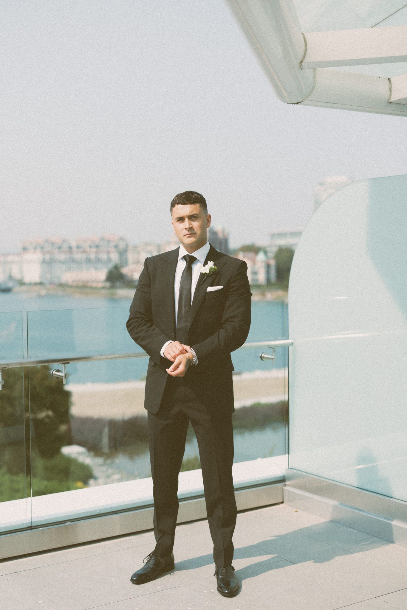 Man in black suit standing on balcony overlooking waterfront view.