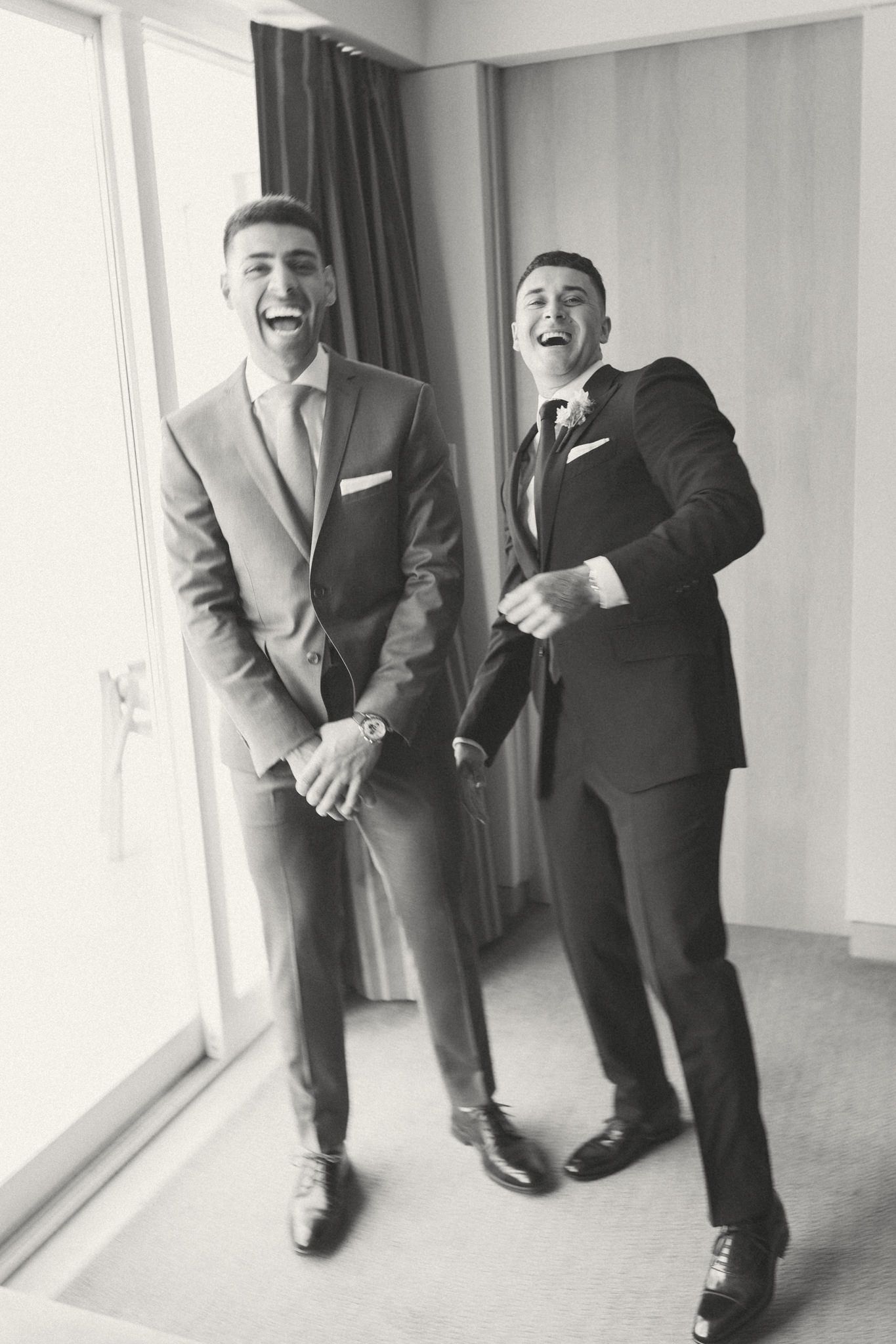 Groomsmen in black suits laughing and posing near window in candid shots.