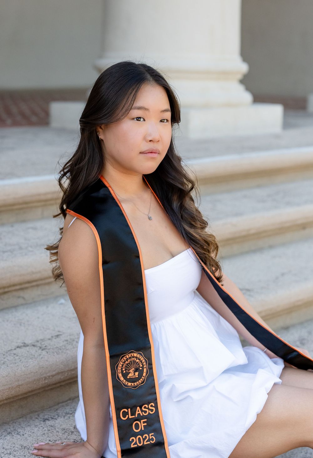 Graduate wearing a black graduation stole with orange text and white dress sitting on stone steps.