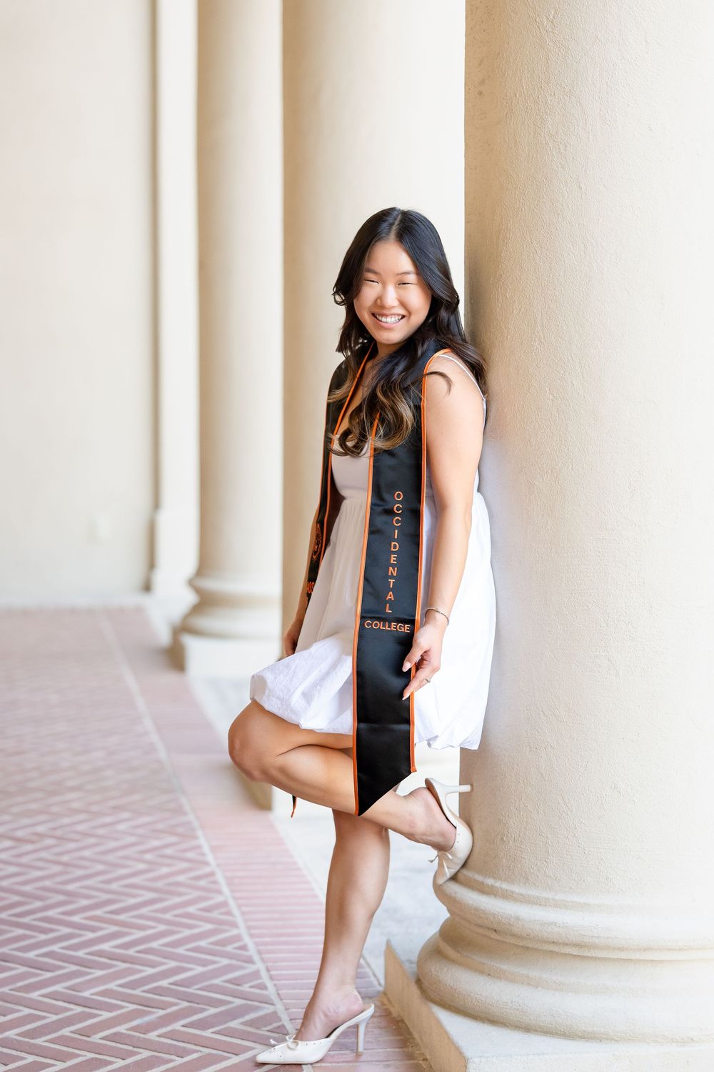 A graduate in a black dress poses with their diploma sash against white columns and brick flooring.