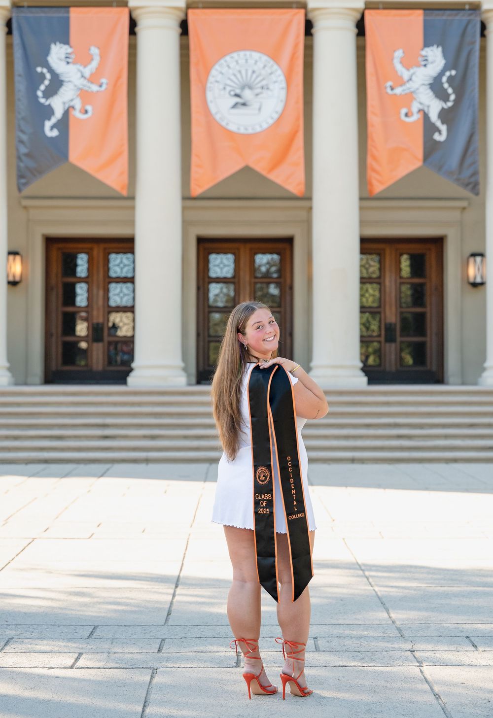 Graduate in white dress and black stole poses in front of columned building with orange banners.