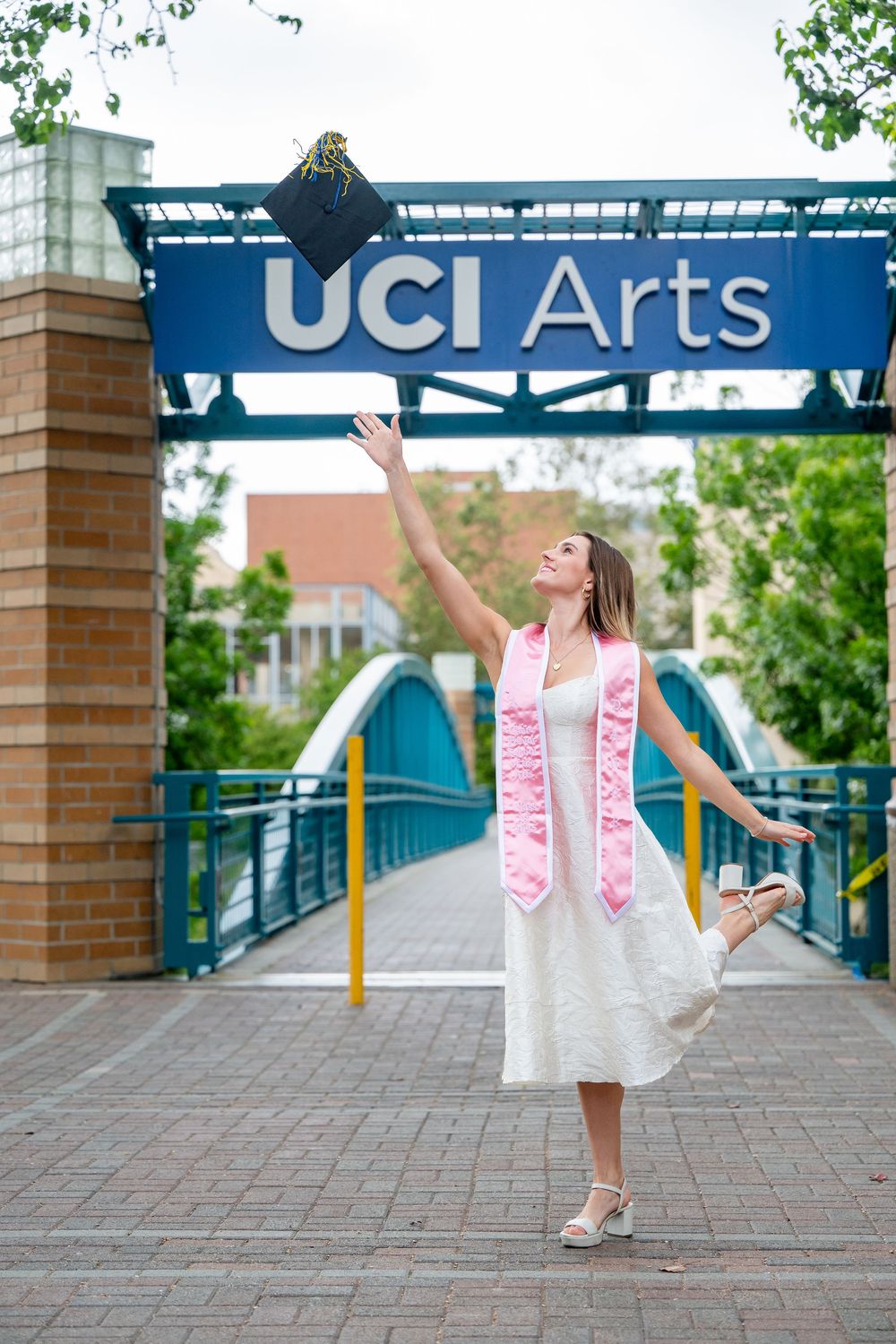 A graduate in a white dress and pink sash celebrates on a campus walkway at UCI Arts with joyful jumping poses.