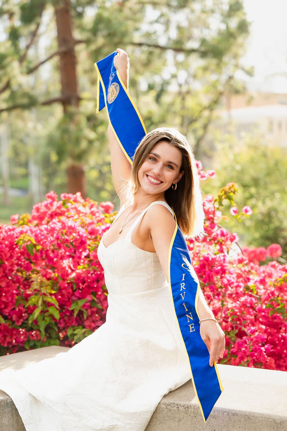 Graduate in white dress poses with blue sash among blooming azalea bushes on sunny spring day.