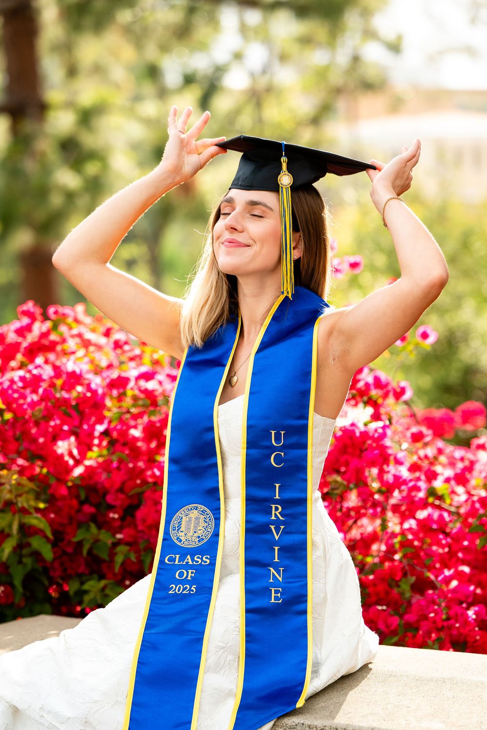 Graduate in blue and white regalia poses joyfully against vibrant red flowers during outdoor graduation photoshoot.
