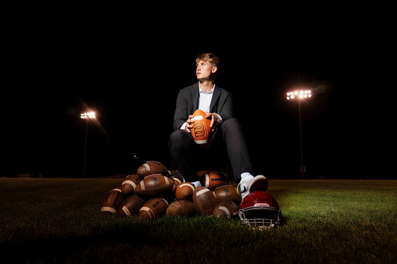 High school football player poses with helmet and ball in front of scoreboard at sunset.