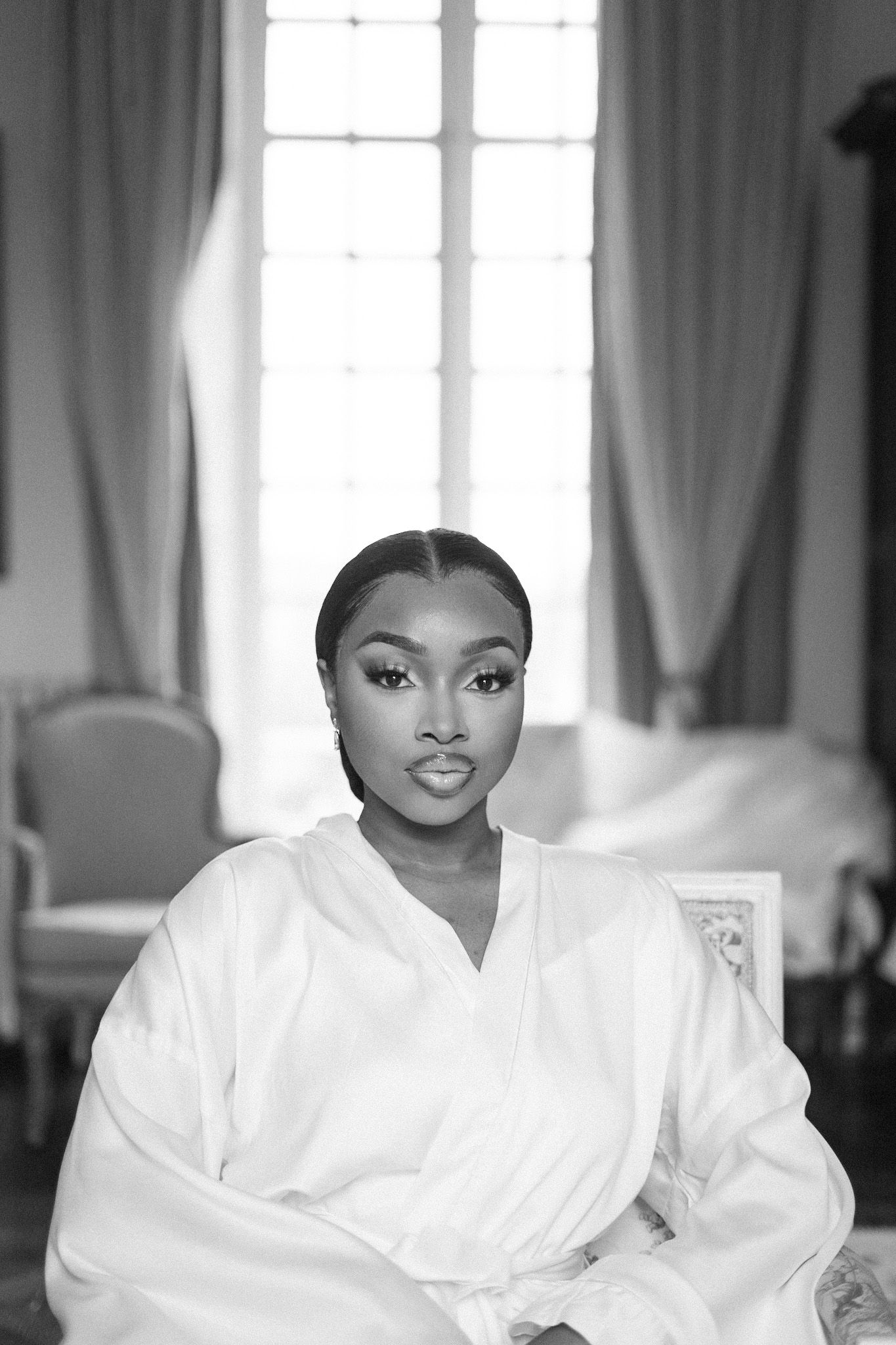 A black and white photograph shows a figure wearing a white bathrobe while sitting in a room with curtained windows.