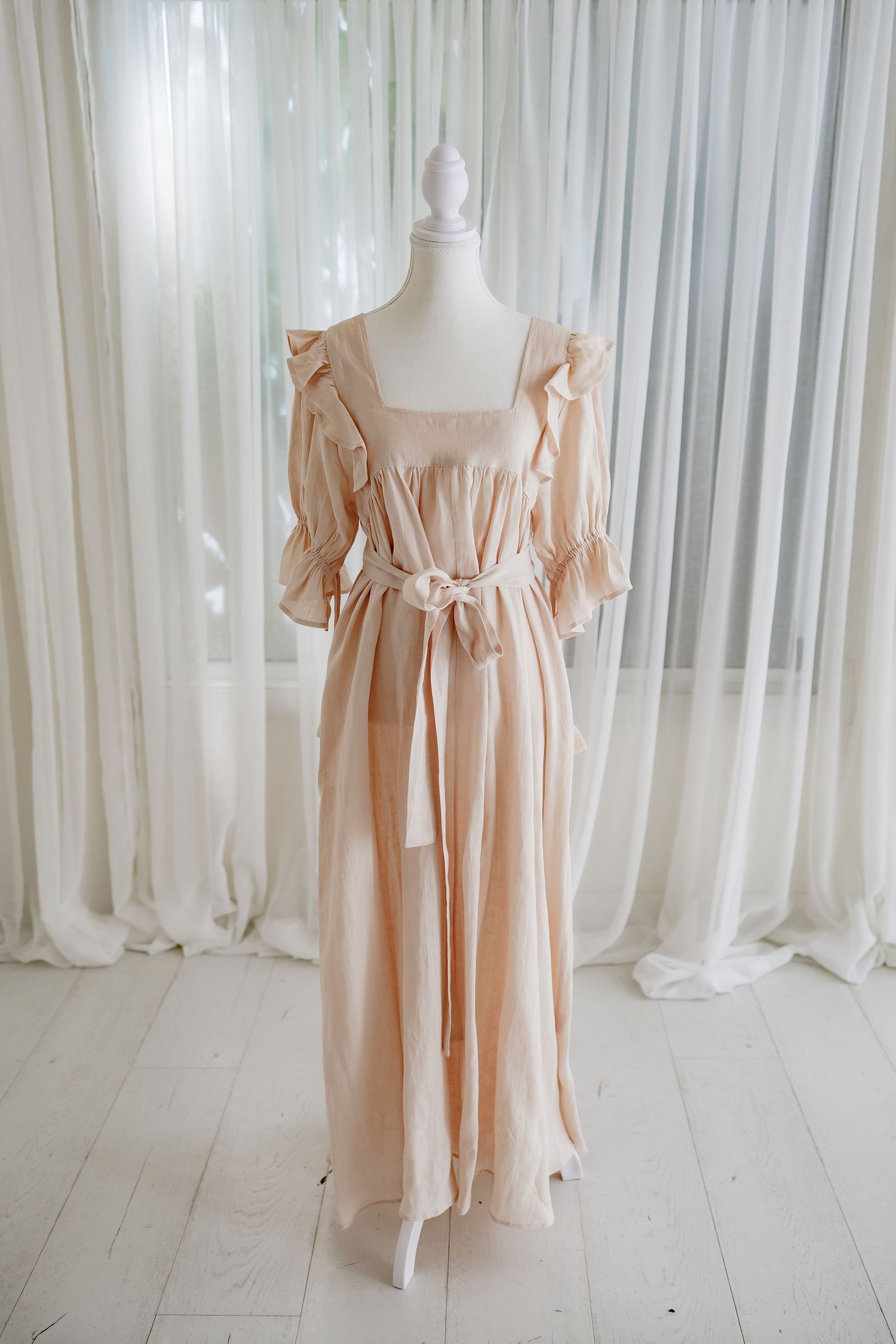 Romantic peach-colored dress with ruffled sleeves and tie waist on mannequin against white curtains.