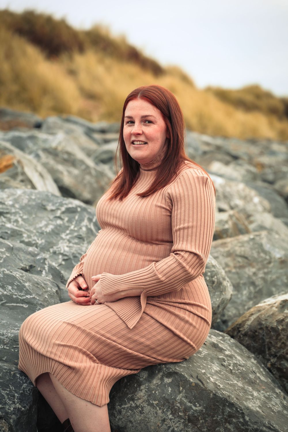 Pregnant woman in beige knit dress smiles while sitting on rocky coastal terrain.