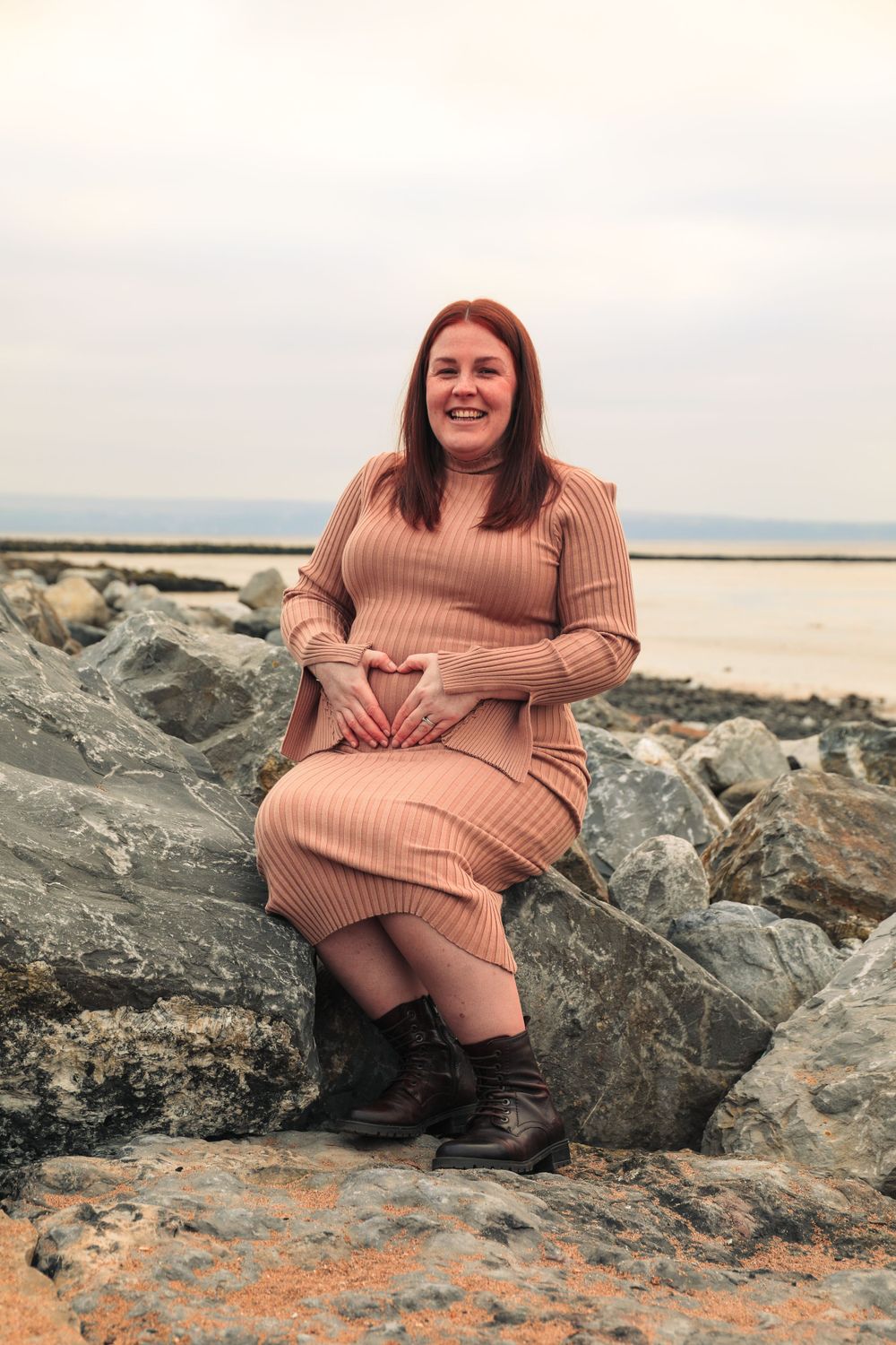 Woman in pink sweater dress sits on coastal rocks near ocean.