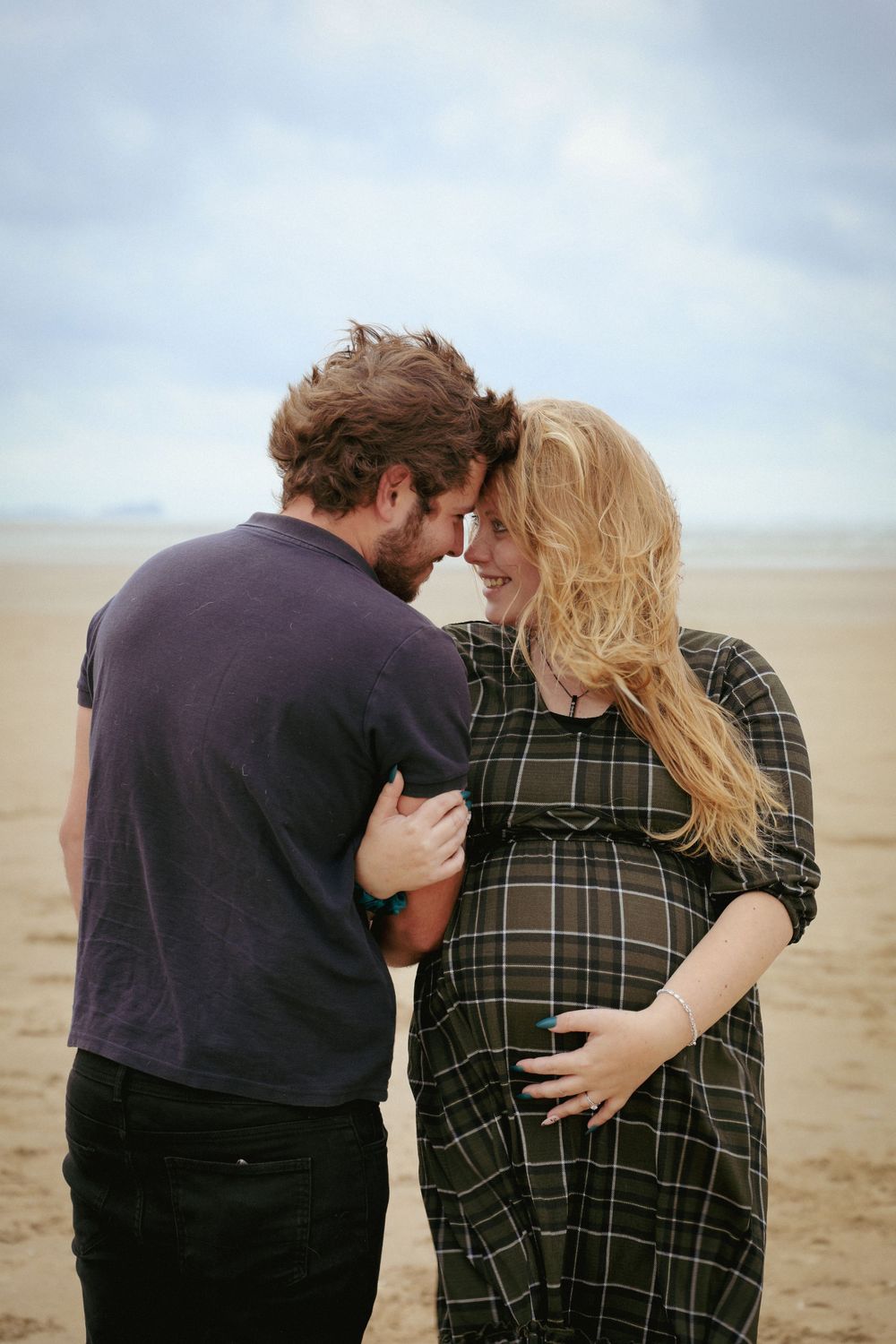 Intimate moment between expecting couple on cloudy beach.
