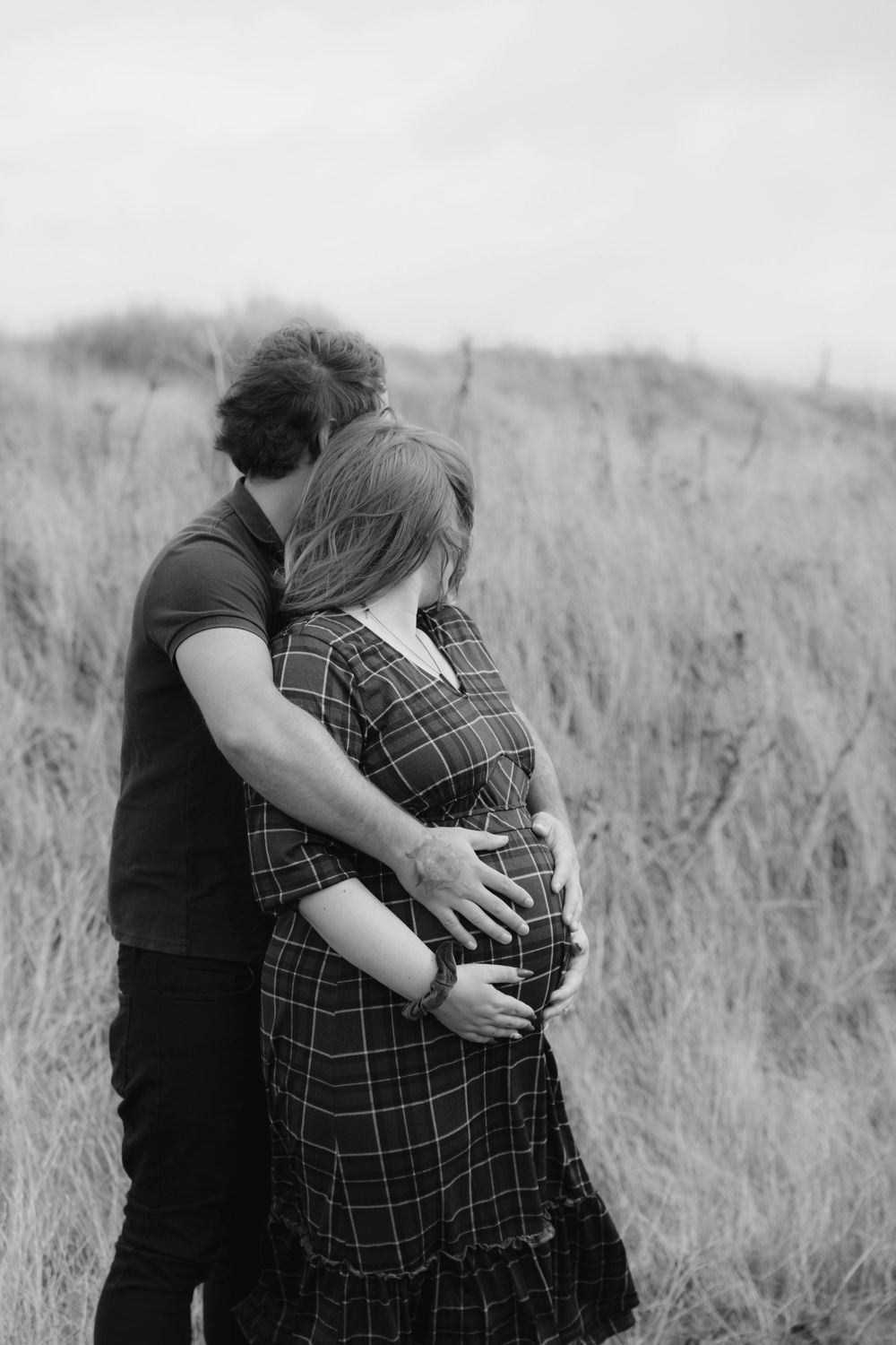 Black and white portrait of an expecting couple embracing in a grassy field.