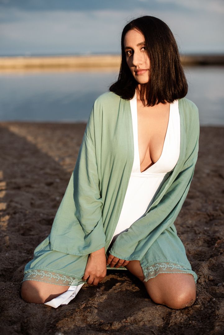 Person in mint green cardigan and white top poses on beach at sunset.