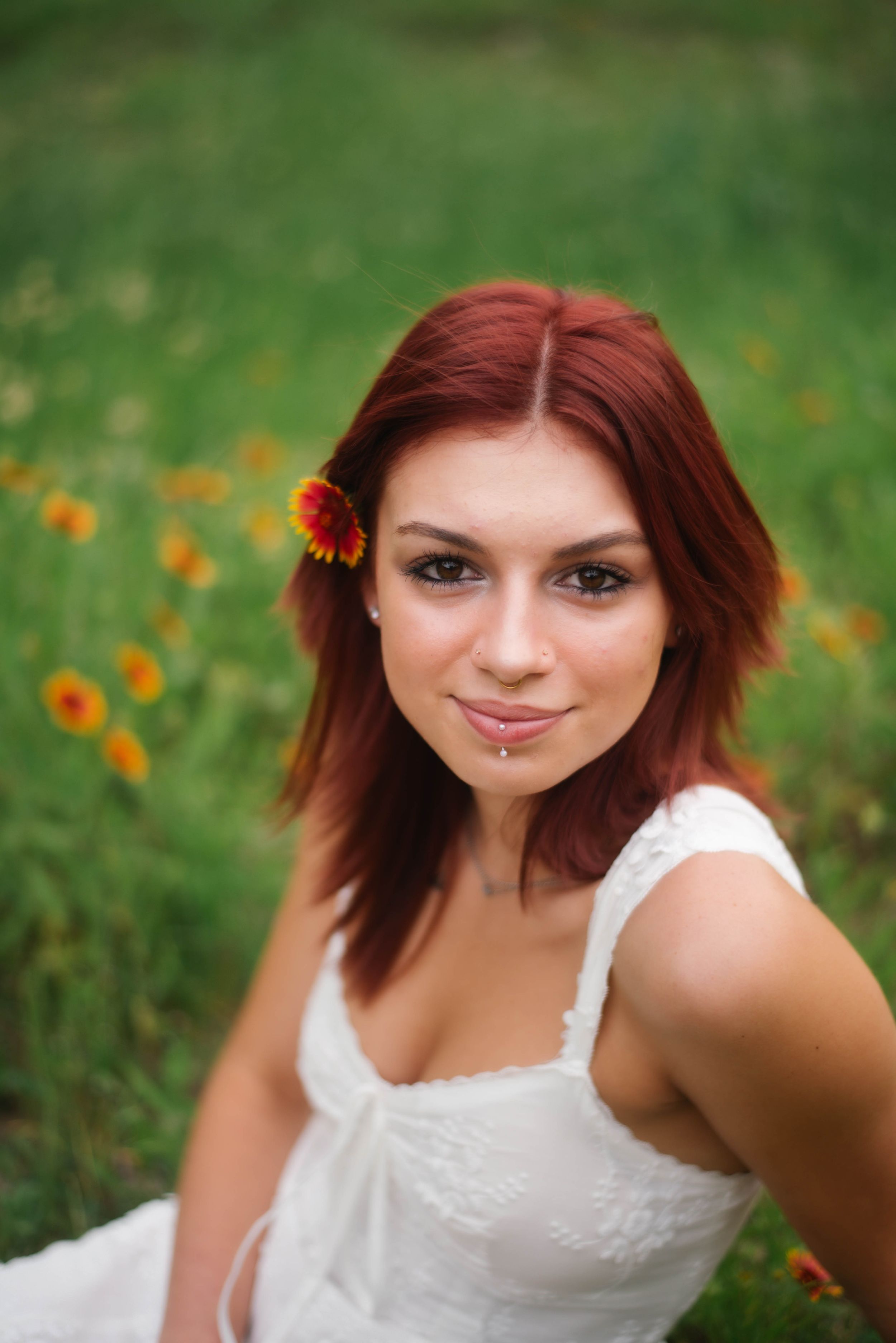 Young woman with red hair and flower accessory smiling in white dress among wildflowers in green field.