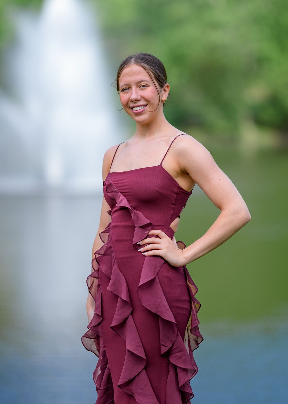 Formal portrait in burgundy ruffled dress against water fountain background.
