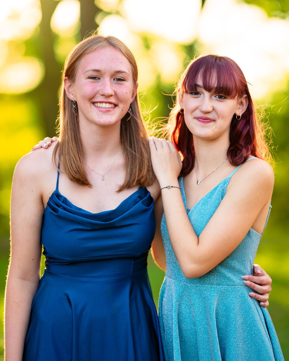 Two friends in elegant blue and turquoise dresses pose together during sunset outdoors.