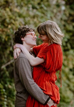 Couple embracing outdoors with one wearing a vibrant red dress and the other in a brown sweater.