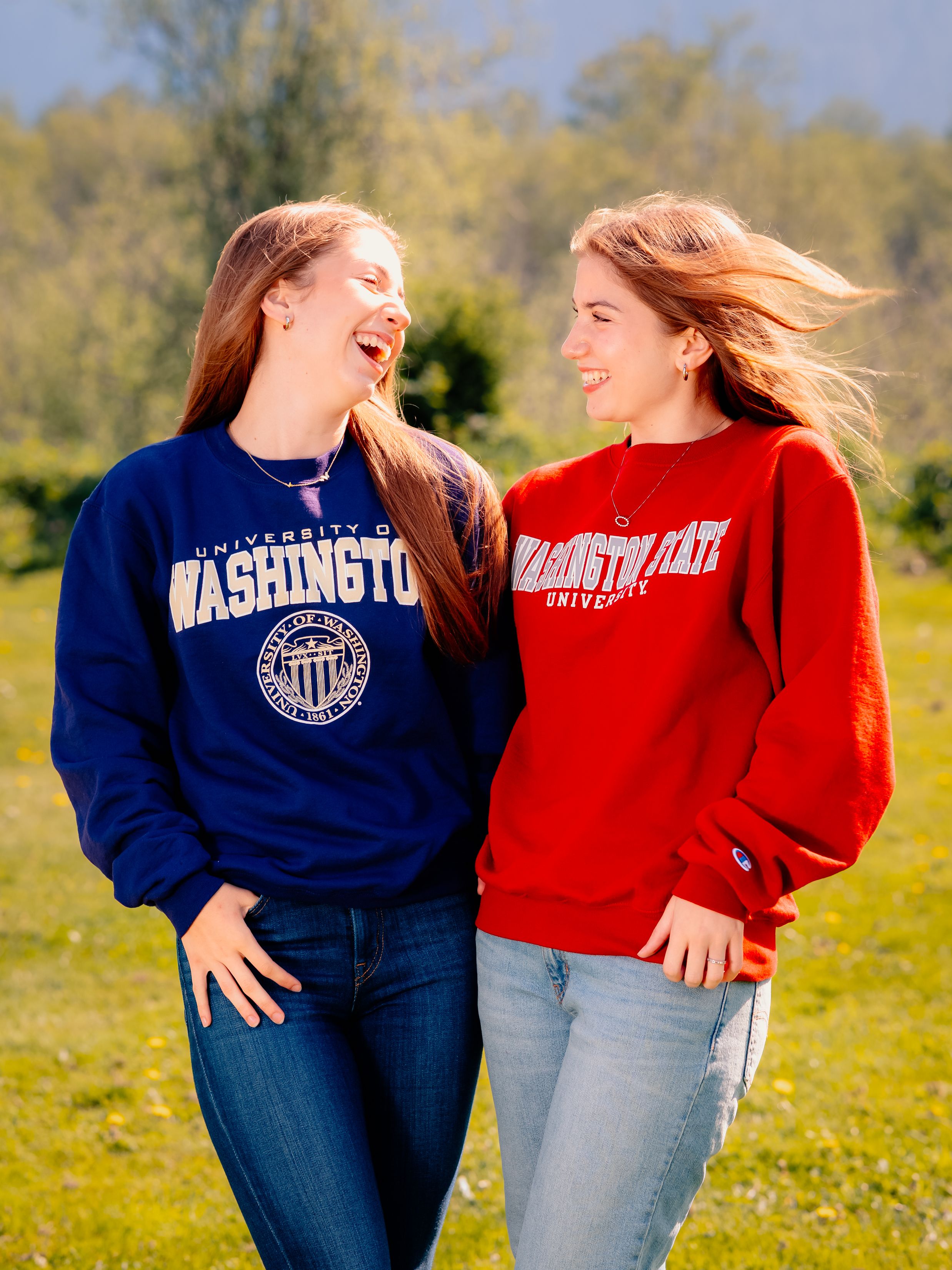 Friends laughing together outdoors wearing university sweatshirts in a sunny field.