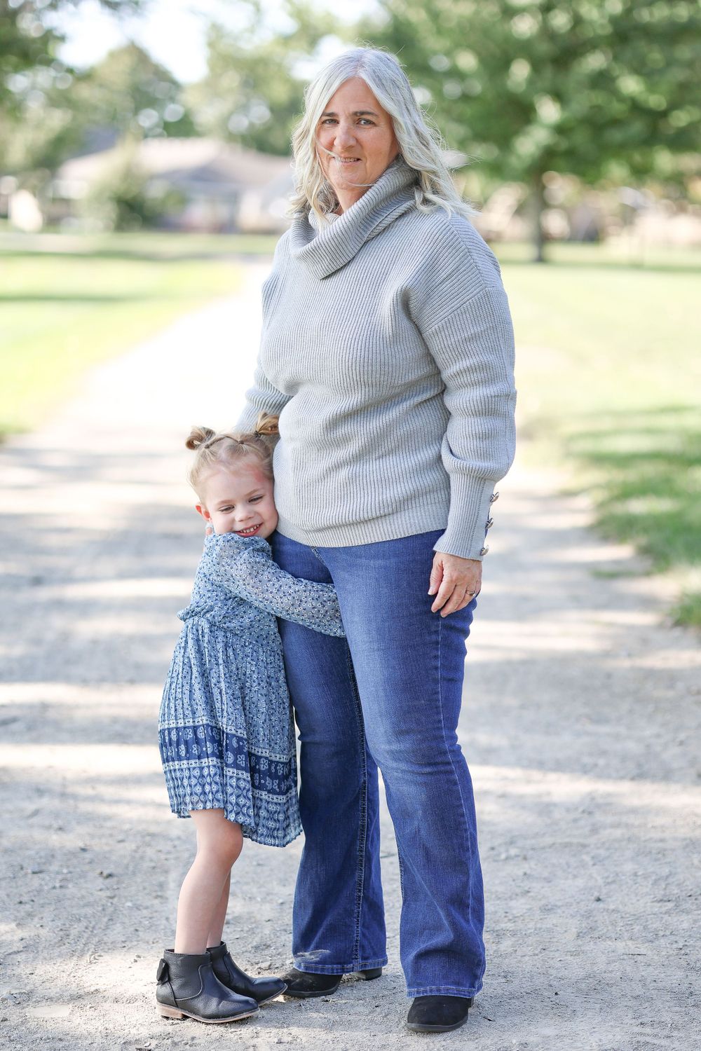 Person in grey sweater stands with child in blue dress on outdoor path.