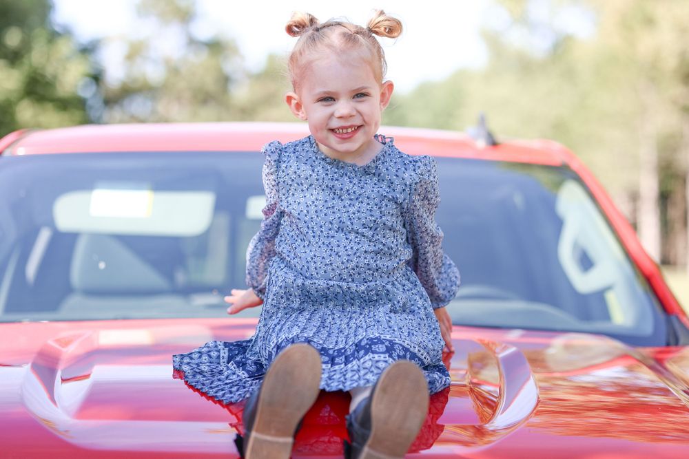 Child in blue dress sitting on red car hood with joyful expression.