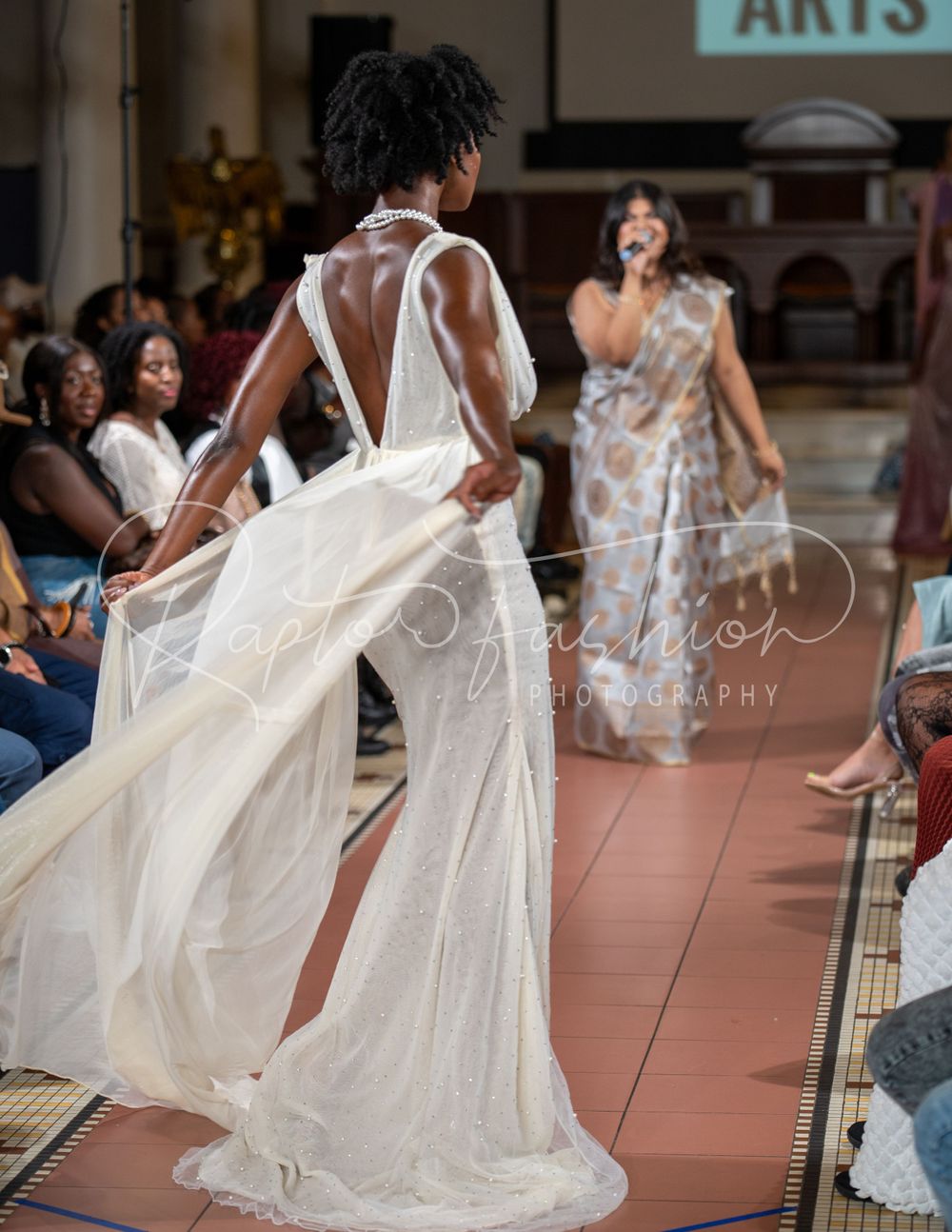 Fashion model in flowing white gown walks runway at arts fashion show.
