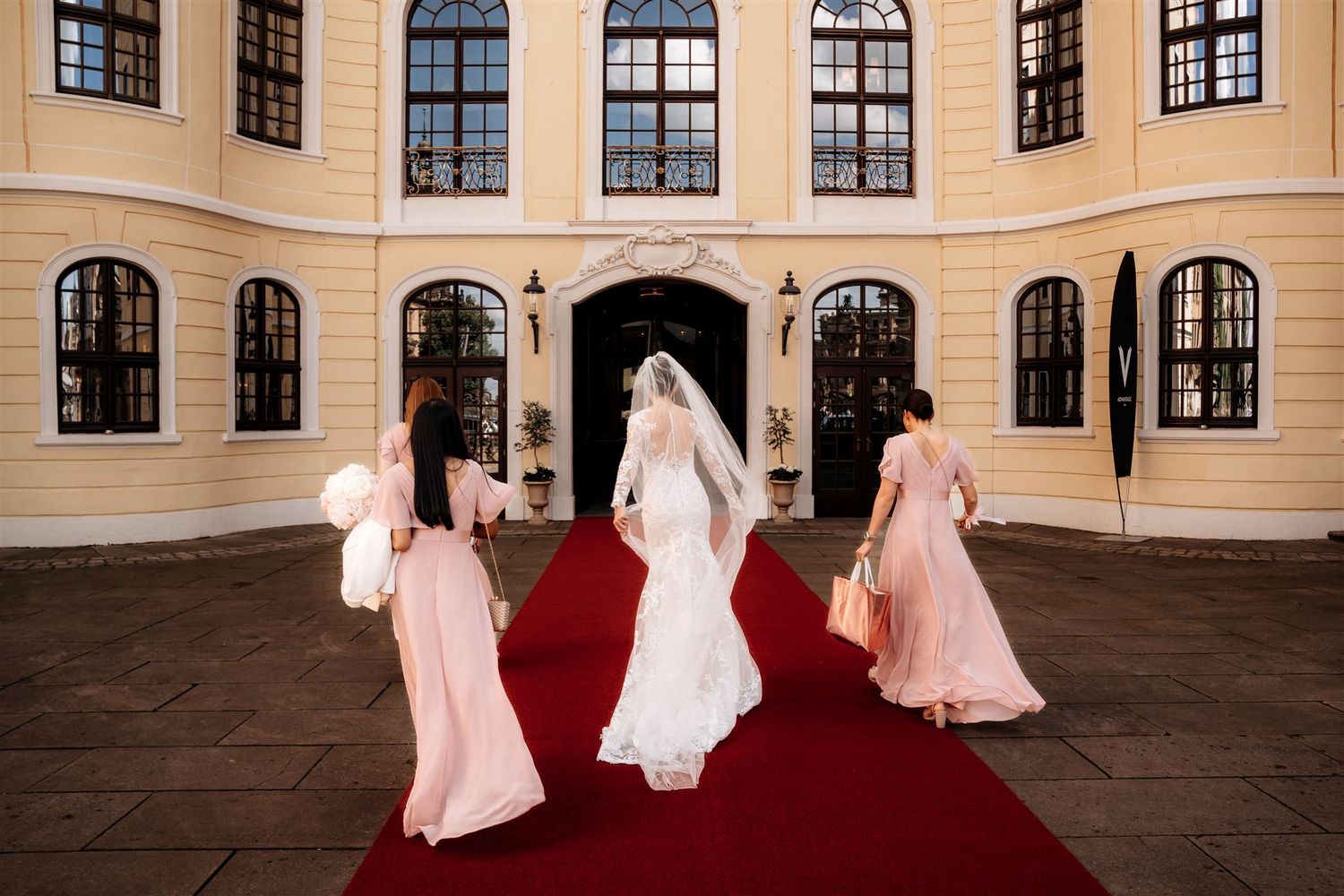 Group walking on red carpet entering elegant historic building with arched windows and cream colored facade.