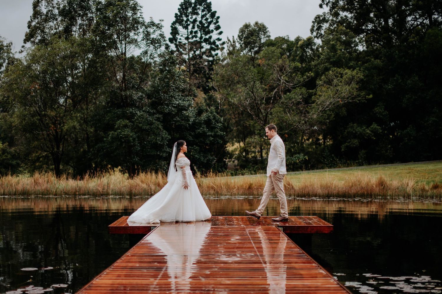 Wedding couple standing on opposite ends of a wooden dock over still water.
