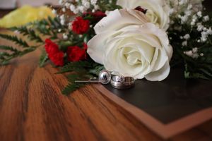 Two silver wedding rings rest on a wooden surface beside a bouquet of white roses and red carnations.