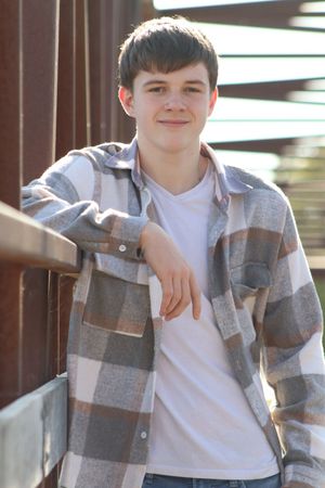 A smiling teenage boy in a plaid flannel shirt leans against a metal railing outdoors in natural light.