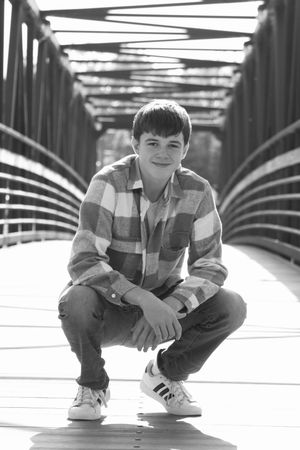 Black and white photo of a teenage boy crouching on a wooden bridge, wearing a plaid shirt and Adidas sneakers.
