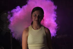 A figure in a light colored tank top stands amid swirling purple smoke effects.