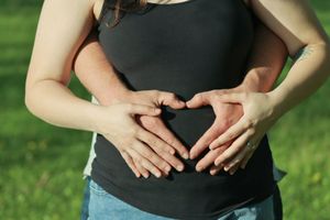 Pregnant woman and partner forming heart shape with hands on baby bump outdoors in sunny green park.