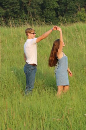 Couple dancing in a lush green meadow, man spinning woman in denim dress, trees in background.