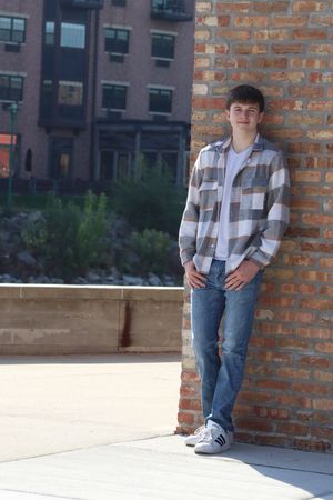 Young man in plaid shirt and jeans leaning against brick wall in urban setting with building background.
