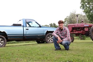 Young man crouching on grass beside a blue pickup truck and red vintage Farmall tractor on a rural farm.