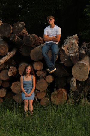 Two young people posing near a large stack of cut logs outdoors, girl sitting on grass, boy perched on top of logs.
