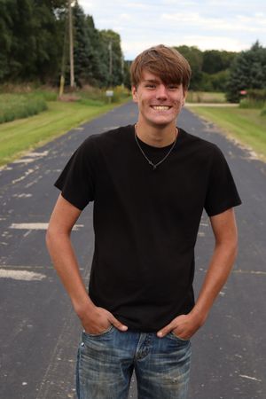 Smiling young man in black t-shirt and jeans standing on a rural road lined with green trees.