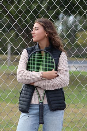 Young woman hugging a green tennis racket, wearing a vest and sweater, standing against a chain-link fence outdoors.