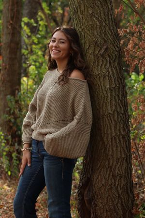 Young woman in beige knit sweater and jeans leaning against a tree, smiling in an autumn forest setting.
