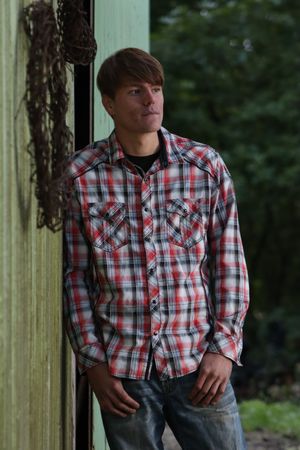 Young man in red plaid shirt leaning against green wall outdoors, looking away thoughtfully.