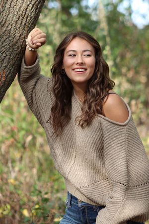 Young woman in beige off-shoulder sweater smiling outdoors, leaning against a tree in a natural fall setting.