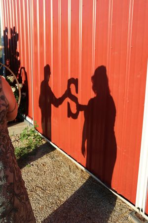 Two people's shadows cast on a bright red corrugated metal barn wall, appearing to share a drink outdoors.