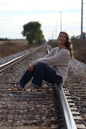 Smiling young woman sitting on railroad tracks in jeans, sweater, and brown Nike sneakers, with trees and sky in background.