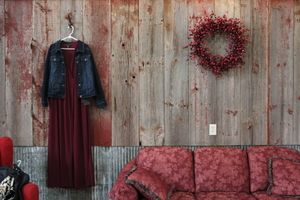A rustic wall display with denim jacket and red dress hanging next to a vintage couch beneath a red berry wreath.